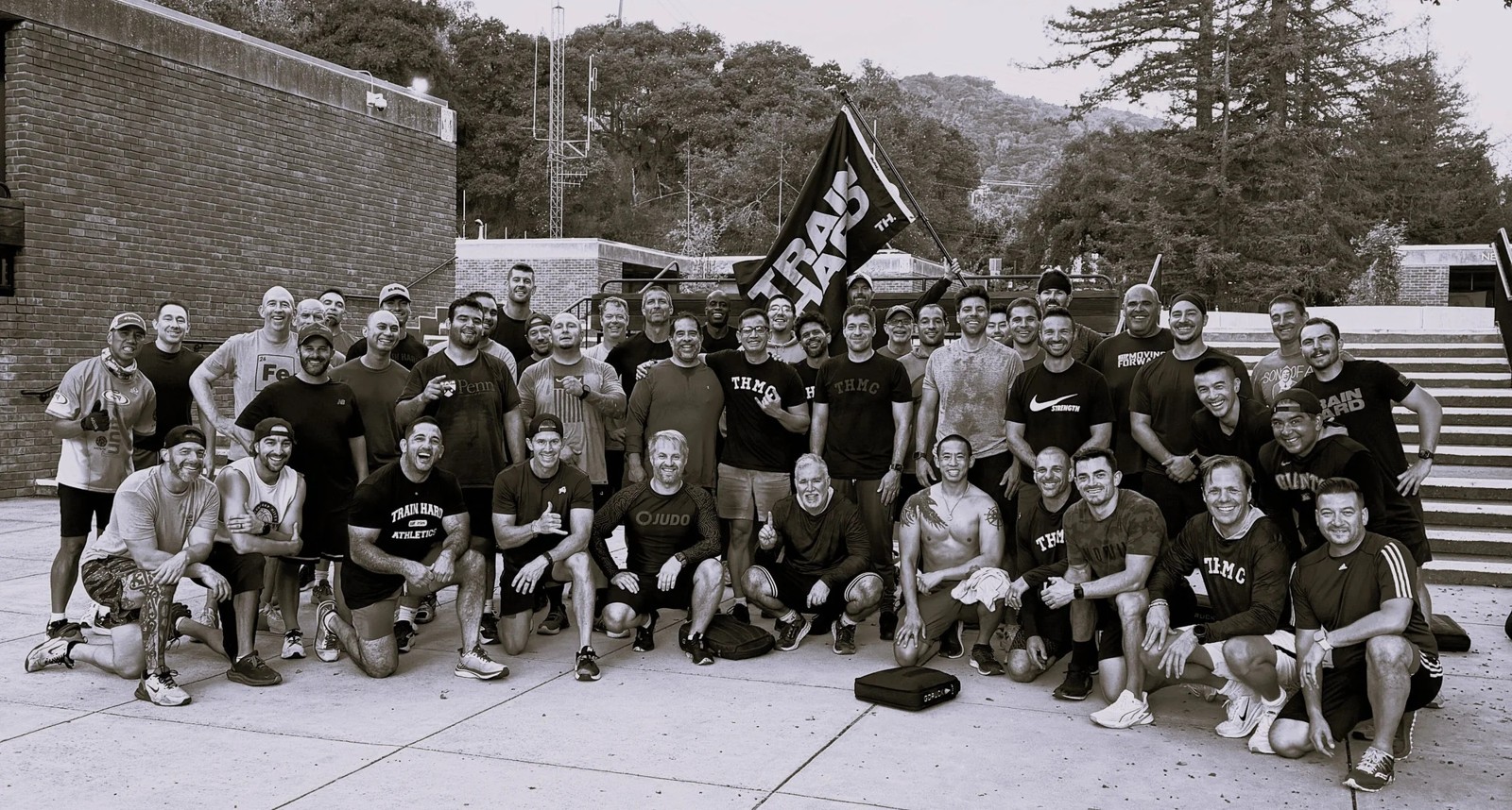 A group of men gathered outdoors beneath a TRAIN HARD flag